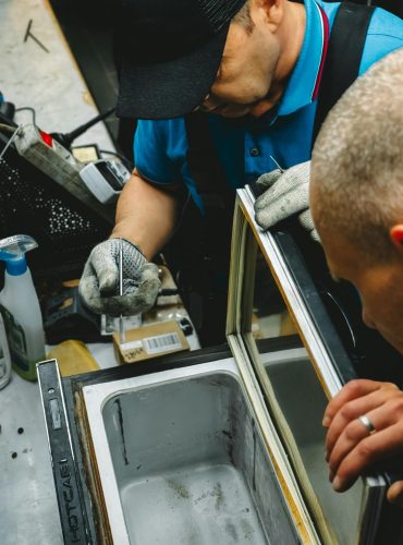 Two technicians work on repairing an industrial freezer in a workshop setting.